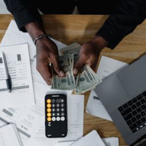 Person counting US dollars, using a calculator and laptop, with financial documents on a wooden desk.