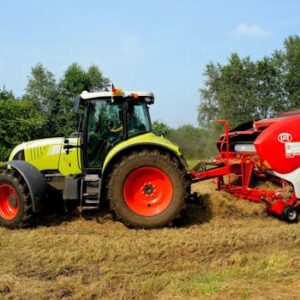 Green tractor and hay baler working on a farm field with trees in the background.